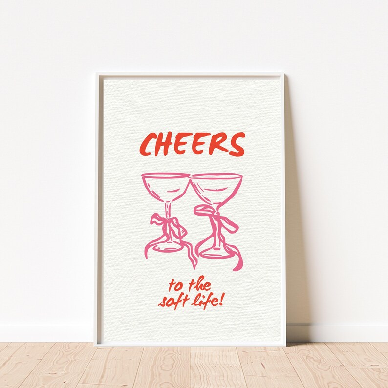 Digital wall art with pink illustrated champagne glasses and red text that says “Cheers to the soft life!” on a textured cream background.