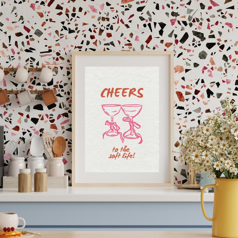 Digital wall art with pink illustrated champagne glasses and red text that says “Cheers to the soft life!” on a textured cream background.