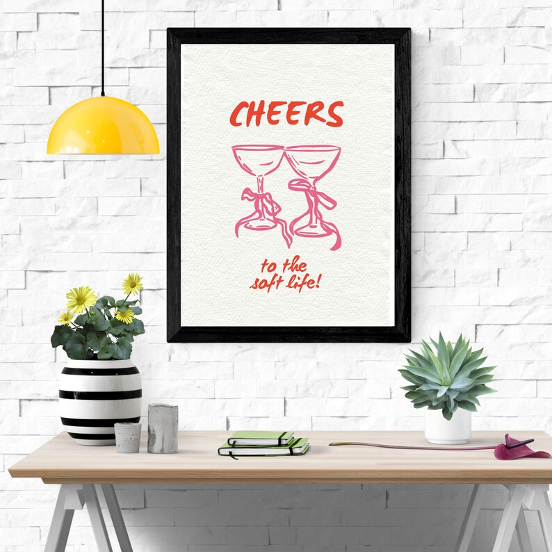 Digital wall art with pink illustrated champagne glasses and red text that says “Cheers to the soft life!” on a textured cream background.