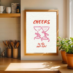 Digital wall art with pink illustrated champagne glasses and red text that says “Cheers to the soft life!” on a textured cream background.