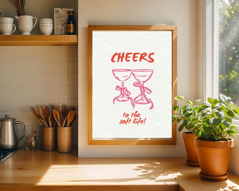 Digital wall art with pink illustrated champagne glasses and red text that says “Cheers to the soft life!” on a textured cream background.