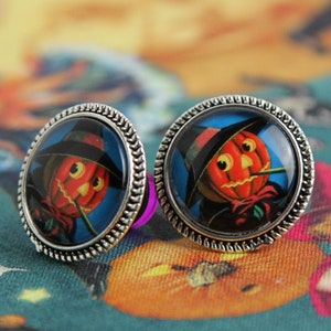 May include: A pair of silver-toned stud earrings. Each earring features a round, black-bordered image of a jack-o'-lantern wearing a black hat against a blue background. The earrings are set against a teal Halloween-themed fabric.