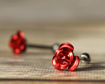 May include: Two red rose-shaped barbell earrings. The earrings have a silver-colored post and a small ball on the back. The rose petals are detailed and the earrings are set against a wooden background.