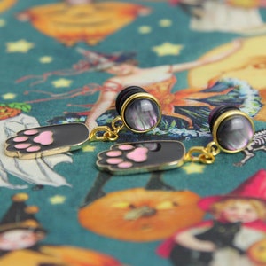 May include: A pair of black and pink cat paw earrings with gold accents and iridescent black and white plugs. The earrings are on a Halloween themed fabric background.