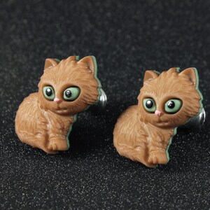 May include: A pair of cat-shaped earrings. Each earring features a brown cat with green eyes and a pink nose. The cat's outline is accented with green. The earrings are set against a dark, speckled background.
