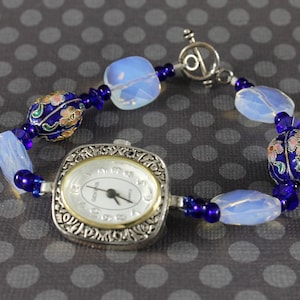 May include: A silver watch with a square face and a blue and white beaded bracelet. The watch face has a white background with black hands and black Roman numeral markings. The bracelet has blue glass beads and white opaque beads with floral designs.