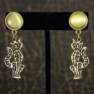 May include: A pair of earrings featuring a light green cabochon top and a dangling gold-colored cat silhouette. The cat design has intricate cut-out details. The earrings are displayed on a black stand against a dark background.