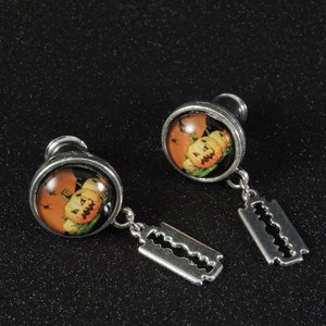 May include: A pair of Halloween-themed earrings. Each earring features a silver-toned plug with a round, orange image of jack-o'-lanterns. A small, silver-toned razor blade charm hangs below each plug, adding a unique touch.