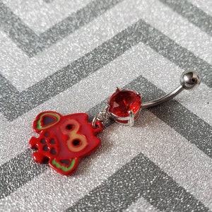 May include: A curved belly button ring with a silver-colored barbell and a red gem. A red charm in the shape of a stylized pig with green and purple accents hangs from the gem.
