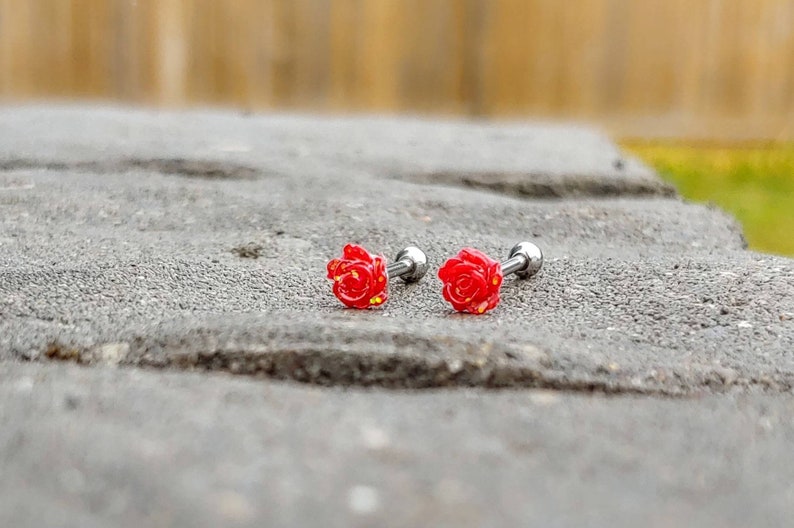 May include: Two red rose flower earrings with silver posts. The earrings are on a gray surface.