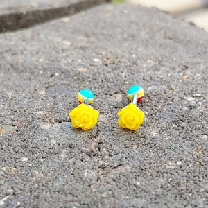 May include: Two yellow rose flower body jewelry pieces with blue, yellow, and red accents. The jewelry is on a gray textured surface.
