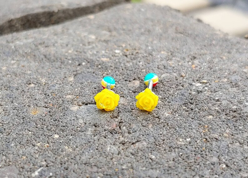 May include: Two yellow rose flower body jewelry pieces with blue, yellow, and red accents. The jewelry is on a gray textured surface.