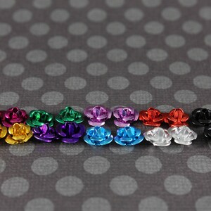 May include: A collection of small, metallic rose-shaped embellishments in various colors, including gold, pink, green, purple, blue, red, silver, and black. The roses are arranged on a gray background with white polka dots.