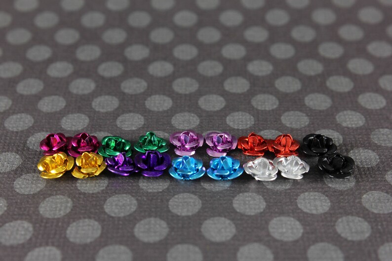 May include: A collection of small, metallic rose-shaped embellishments in various colors, including gold, pink, green, purple, blue, red, silver, and black. The roses are arranged on a gray background with white polka dots.