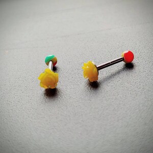 May include: Two yellow rose-shaped barbell jewelry pieces. One barbell has a red ball on one end and a yellow rose on the other. The other barbell has a green, red, and yellow ball on one end and a yellow rose on the other.