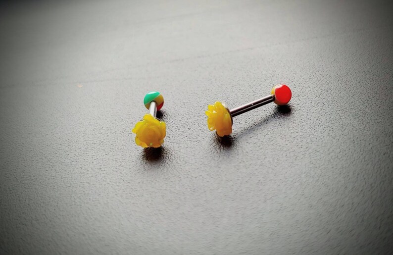 May include: Two yellow rose-shaped barbell jewelry pieces. One barbell has a red ball on one end and a yellow rose on the other. The other barbell has a green, red, and yellow ball on one end and a yellow rose on the other.