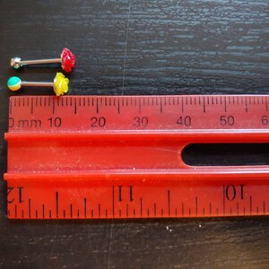 May include: Two pairs of body jewelry with small, colorful flower accents. The jewelry is measured against a ruler with markings in millimeters.