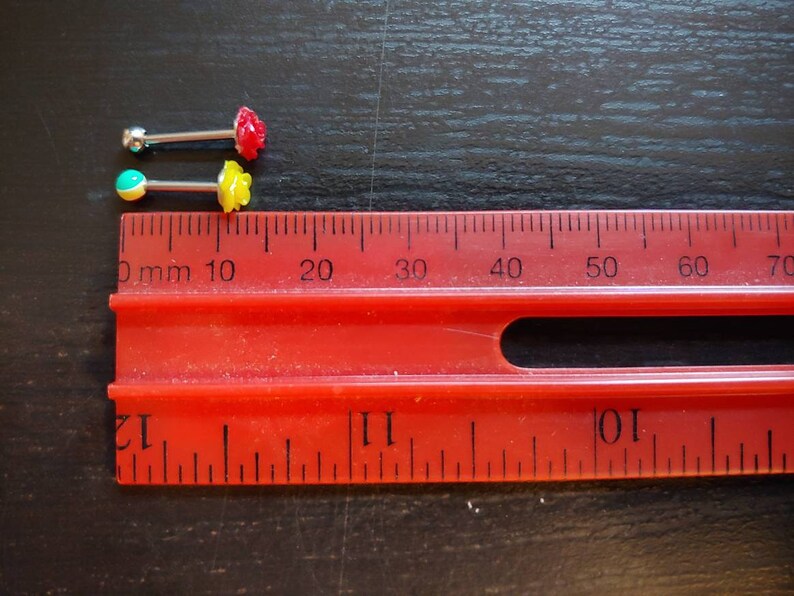 May include: Two pairs of body jewelry with small, colorful flower accents. The jewelry is measured against a ruler with markings in millimeters.