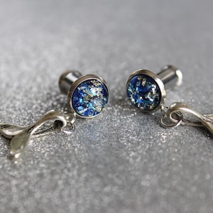 May include: A pair of silver earrings with blue and gold glitter accents. Each earring features a small, round, blue and gold glitter charm with a silver backing. The earrings are attached to a silver chain with a small, silver fishtail charm.