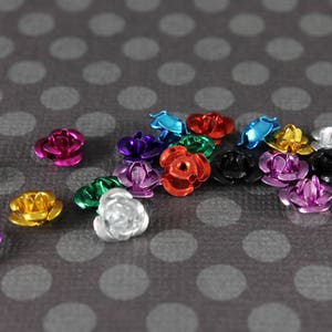 May include: A collection of small, metallic rose-shaped embellishments in various colors, including pink, purple, gold, silver, red, and blue. The decorative flowers are scattered on a gray background with white polka dots.