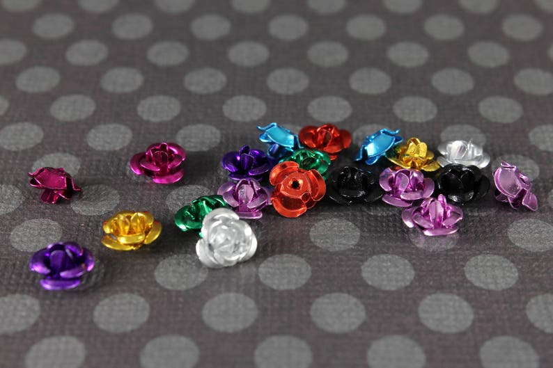 May include: A collection of small, metallic rose-shaped embellishments in various colors, including pink, purple, gold, silver, red, and blue. The decorative flowers are scattered on a gray background with white polka dots.