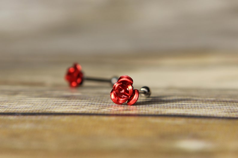 May include: Two red rose-shaped barbell earrings. The earrings have a silver-colored post and a small ball on the back. The rose petals are detailed and the earrings are set against a wooden background.
