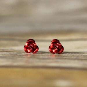 May include: A pair of red rose-shaped stud earrings. The earrings are made of a shiny, metallic material and are set against a blurred wooden background. The earrings are a classic floral design.