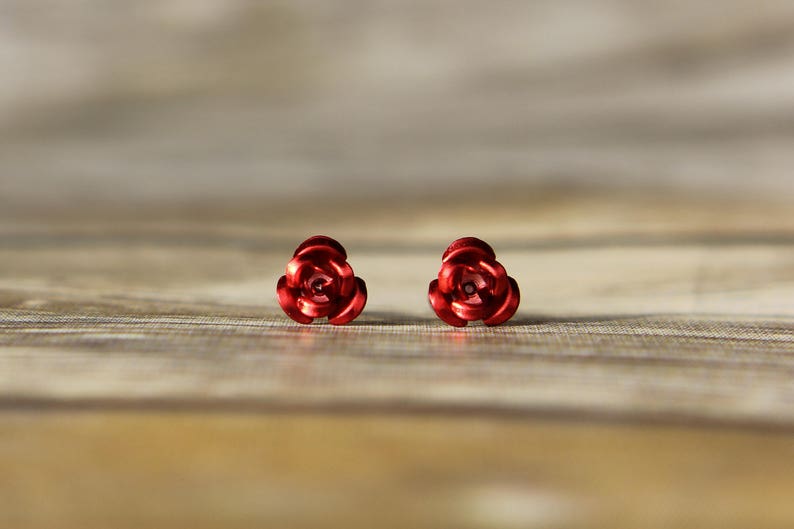 May include: A pair of red rose-shaped stud earrings. The earrings are made of a shiny, metallic material and are set against a blurred wooden background. The earrings are a classic floral design.
