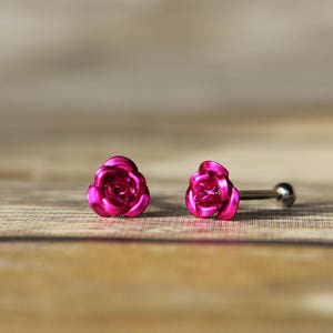 May include: A pair of bright pink rose-shaped stud earrings. The earrings are made of a shiny metal and have a post and ball closure. The earrings are set against a blurred wooden background.