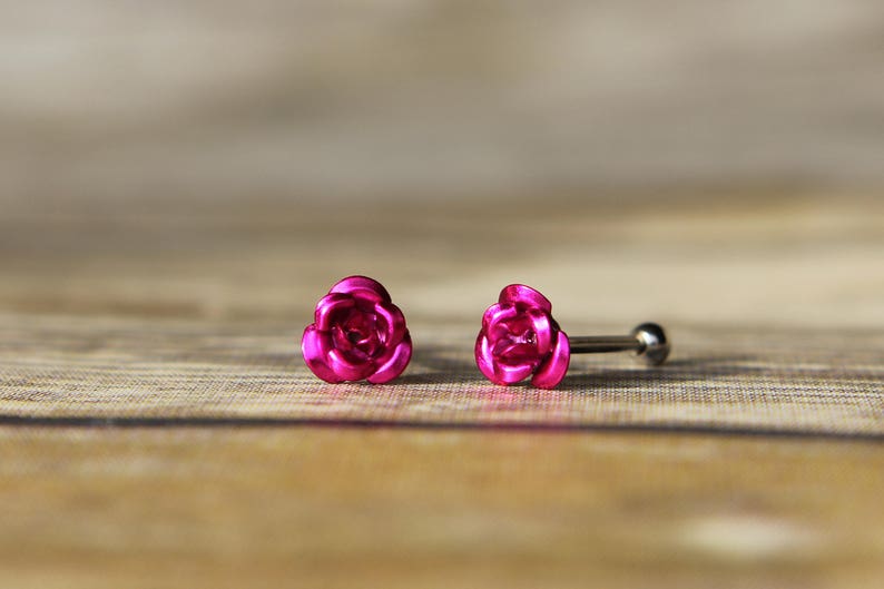 May include: A pair of bright pink rose-shaped stud earrings. The earrings are made of a shiny metal and have a post and ball closure. The earrings are set against a blurred wooden background.