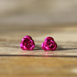 May include: A pair of bright pink rose-shaped stud earrings. The earrings are made of a shiny material and are set against a wooden background. The earrings are a close-up shot.