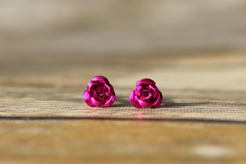 May include: A pair of bright pink rose-shaped stud earrings. The earrings are made of a shiny material and are set against a wooden background. The earrings are a close-up shot.