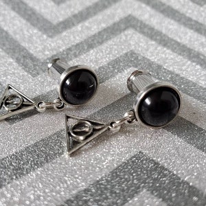 May include: A pair of silver ear gauges with black stone plugs and a silver triangle charm with a circle in the center. The charm is attached to the plug with a silver chain.