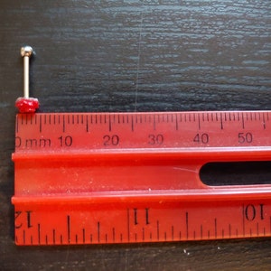 May include: A close-up of a red and silver barbell piercing with a small red gem on the end. The piercing is measured against a red ruler with black markings in millimeters.