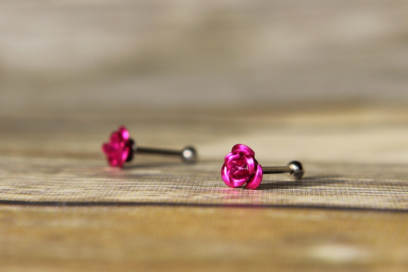 May include: Two pieces of jewelry, each featuring a bright pink rose design. The rose is the focal point of the jewelry, with a silver-colored post and ball backing. The earrings are set against a wooden background.
