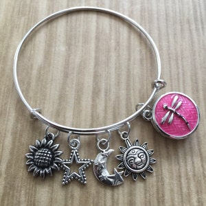 May include: A silver bangle bracelet with charms including a sunflower, a star, a crescent moon, a sun with a face, and a pink dragonfly charm.