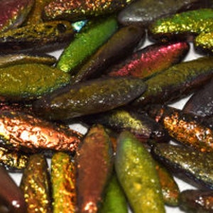 May include: A pile of iridescent glass beads in shades of green, gold, and red. The beads are long and narrow, with a slightly irregular shape.