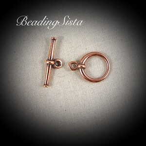 May include: A pair of copper-colored toggle clasps for jewelry making. The clasp on the left is a toggle with a loop on the end. The clasp on the right is a ring.