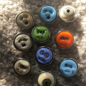 May include: A collection of 10 colorful glass buttons with various cartoon faces and eyes. The buttons are round and have a silver metal rim.