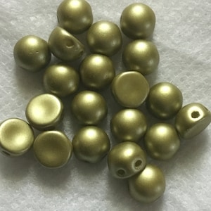 May include: A group of 18 gold-colored round cabochon beads with a hole in the top. The beads are arranged in a pile on a white surface.