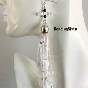 May include: White tassel earrings with a silver bead and a clear crystal with a black bead. The earrings are hanging from a silver hook.