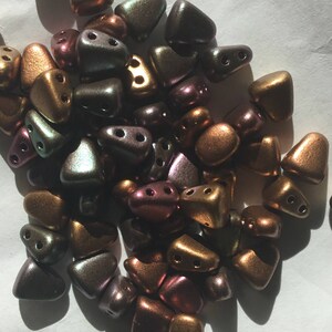 May include: A pile of small, iridescent, triangular beads in shades of gold, brown, and purple. The beads have two holes each and are scattered on a white surface.
