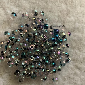 May include: A pile of iridescent glass beads with a clear, transparent appearance. The beads have a subtle sheen of blue, purple, and green, and are scattered on a light gray, textured surface. The beads are small and round.