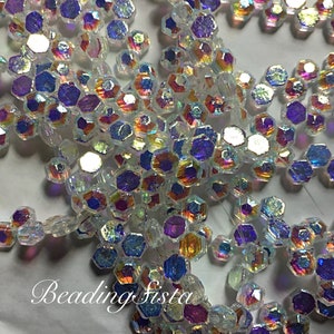 May include: A close-up of iridescent clear glass beads with a hexagonal shape. The beads have a rainbow effect with shades of blue, purple, and orange.