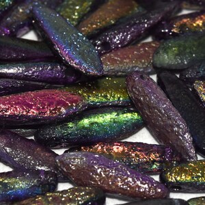 May include: A collection of iridescent glass beads in various colors, including purple, green, and gold. The beads are oblong in shape and have a textured surface, reflecting light. These beads are suitable for jewelry making and crafts.