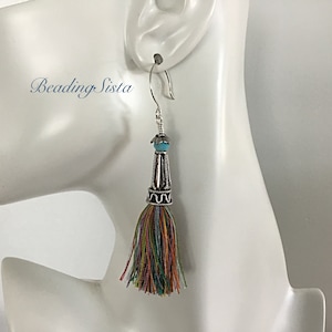 May include: Silver dangle earrings with a turquoise bead and a colorful tassel. The tassel is made of multiple strands of yarn in shades of red, orange, yellow, green, blue, and purple.