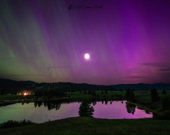 May include: A scenic landscape photograph captures the aurora borealis over a tranquil lake. The sky is a vibrant mix of purple, green, and pink hues, with a full moon and stars. The lake reflects the colorful sky, with dark trees and hills in the background.
