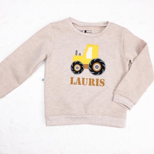 Puede incluir: Una sudadera beige de manga larga con tejido de punto gofrado texturizado. El frente presenta un gráfico de un tractor amarillo con neumáticos negros y el nombre LAURIS impreso en letras mayúsculas marrones debajo del gráfico.
