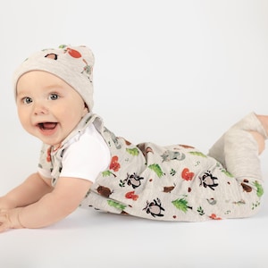 May include: A smiling baby wearing a light gray hat and a matching romper with a whimsical forest animal print. The outfit includes a white short-sleeved shirt underneath and light gray leggings. The baby is lying on their stomach.