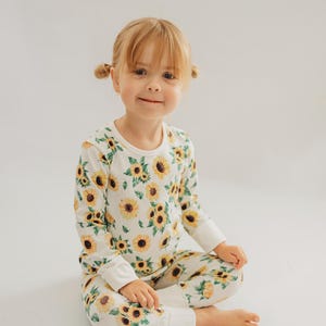 Pode incluir: Um conjunto de pijama de duas peças para crianças. A parte de cima de manga comprida e as calças são brancas com um padrão repetido de girassóis amarelos e folhas verdes. A criança está sentada de pernas cruzadas numa superfície branca.